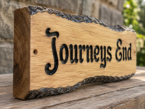 A rustic live-edge wooden sign with the phrase 'Journeys End' carved in black lettering, displayed on a weathered wooden surface with a blurred stone wall and greenery in the background.