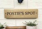 Wooden sign with 'POSTIE'S SPOT' text on a white wall with plants below.